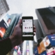 Image shows a hand holding an iphone open on the Instagram app. The phone is being held up to the sky with skyscrapers and colourful advertisements in the background.